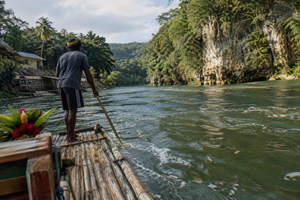 A bamboo raft captain standing at the rear of a raft poling along a wide quiet river
