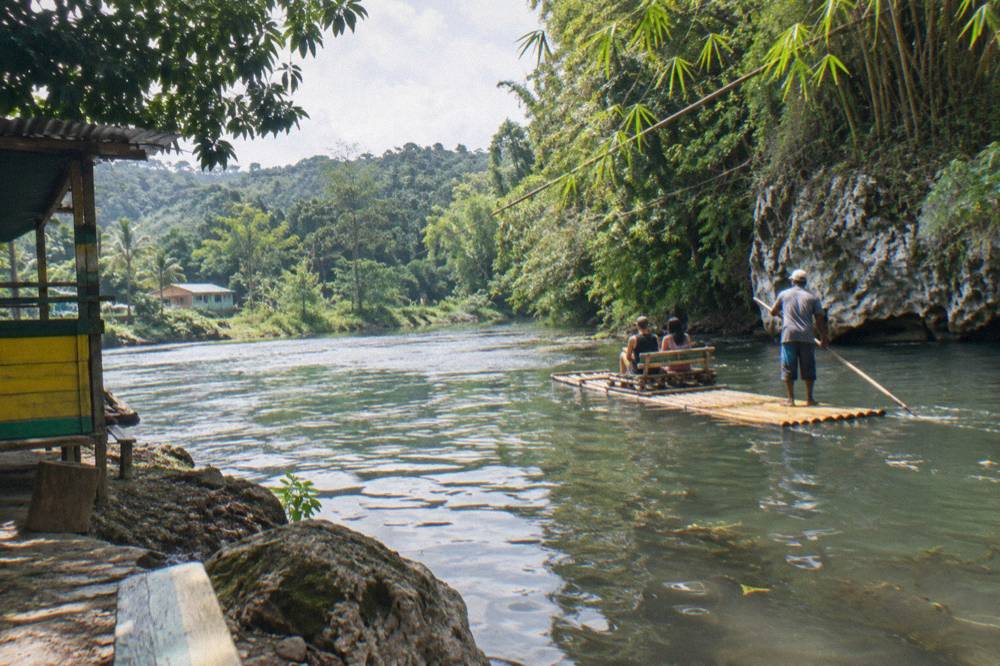 A raft captain poling a bamboo raft along a slow green river bend