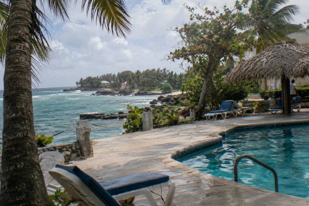A low white resort building with a shaded pool terrace beside a calm Caribbean cove